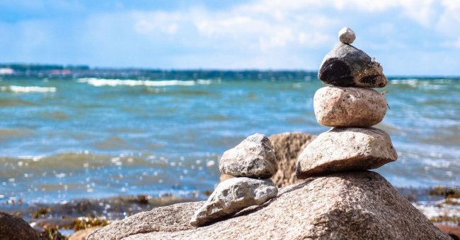 Übereinander gestapelte kleine Steine am Meer. Ganz unten liegt ein größerer Stein. Links liegen zwei kleine Steine neben dem Stapel. Das Meer ist leicht türkis und hat leichte Wellen. Es sind Schaumkronen zu erkennen.
