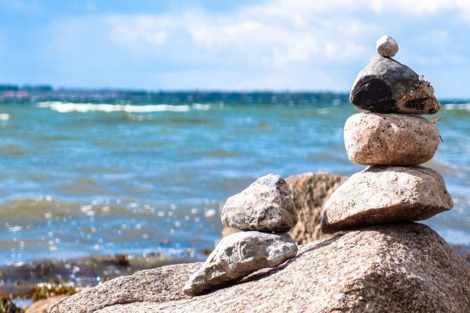 Übereinander gestapelte kleine Steine am Meer. Ganz unten liegt ein größerer Stein. Links liegen zwei kleine Steine neben dem Stapel. Das Meer ist leicht türkis und hat leichte Wellen. Es sind Schaumkronen zu erkennen.