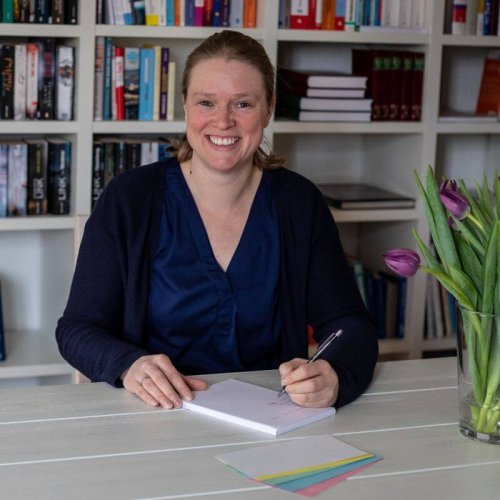 Auf diesem Foto bin ich abgebildet, wie ich etwas auf einen Block schreibe. Ich lächle. Ich trage eine blaue Bluse und eine blaue Strickjacke und sitze an einem weißen Tisch.