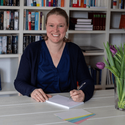 Auf diesem Foto sitze ich an einem weißen Tisch und lächle in die Kamera. Ich schreibe etwas auf einen Block, habe in der linken Hand einen Stift. Vor mir liegen verschiedenfarbige Moderationskarten, rechts von mir (aus deiner Sicht) stehen lilafarbene Tulpen.