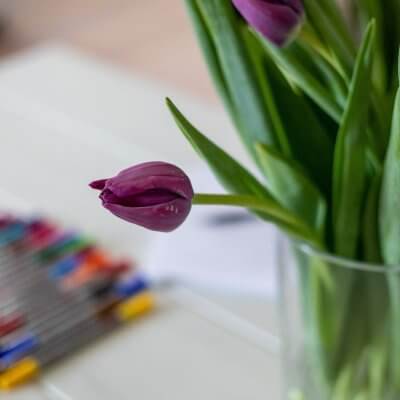 Es sind bunte Filzstifte und lilafarbene Tulpen abgebildet. Beides liegt und steht auf einem weißen Tisch.