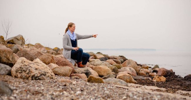 Ich sitze an der Ostsee, auf Steinen und zeige auf das Wasser. Das Wasser ist ganz glatt, da es windstill ist.