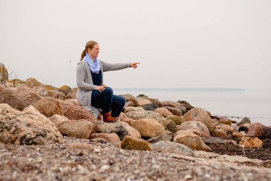 Ich sitze an der Ostsee, auf Steinen und zeige auf das Wasser. Das Wasser ist ganz glatt, da es windstill ist.