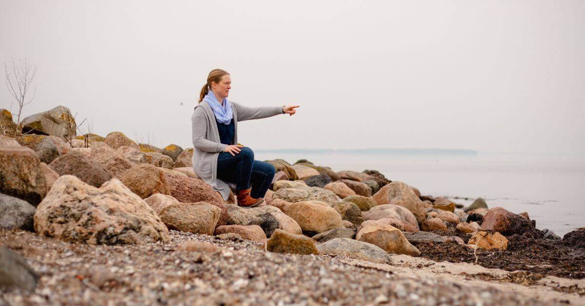 Ich sitze an der Ostsee, auf Steinen und zeige auf das Wasser. Das Wasser ist ganz glatt, da es windstill ist.