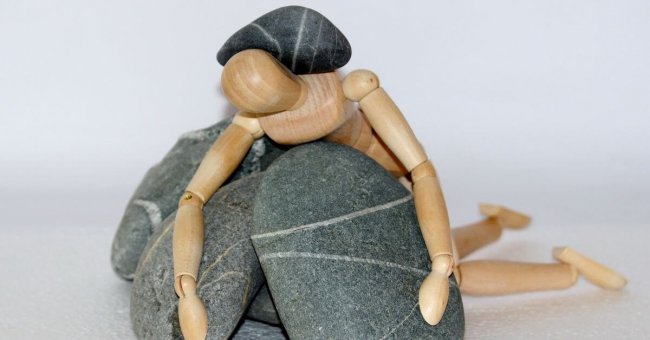 Das Bild zeigt ein Holzmännchen, das auf Steinen liegt und auf dem Körper ebenfalls ein Stein liegen hat.