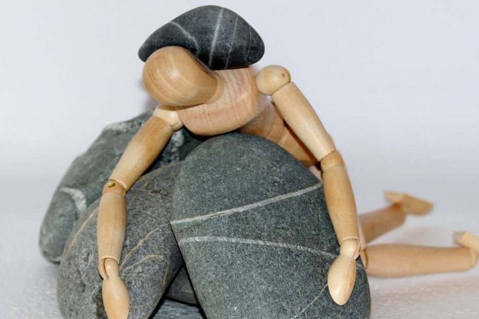 Das Bild zeigt ein Holzmännchen, das auf Steinen liegt und auf dem Körper ebenfalls ein Stein liegen hat.