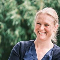 Ich stehe vor dem Bambusbusch und lache in die Kamera. Ich trage eine blaue Bluse und eine blaue Strickjacke. Mein Haar ist zu einem Knoten gebunden. Mein Haar ist dunkelblond.