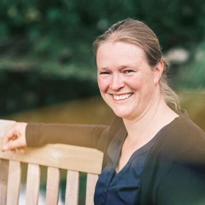 Auf dem Bild bin ich, sitzend auf einer Gartenbank zu ereannen. Mein rechter Arm liegt auf der Rückenlehne. Die Bank ist holzfarbend. Ich habe dunkelblonde Haare und ein strahlendes Lächeln. Ich trage eine blaue Bluse und eine blaue Strickjacke.