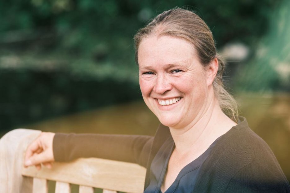 Ich sitze auf einer Gartenbank vor einen großen Baum. Es ist Sommer, der Baum ist dunkelgrün belaubt. Das Foto zeigt mich scharf, den Hintergrund verschwommen. Ich lache, habe zusammengebundene Haare. Dunkelblond. Ich trage eine blaue Bluse und Strickjacke. Mein rechter Arm liegt auf der Rückenlehne der Bank.