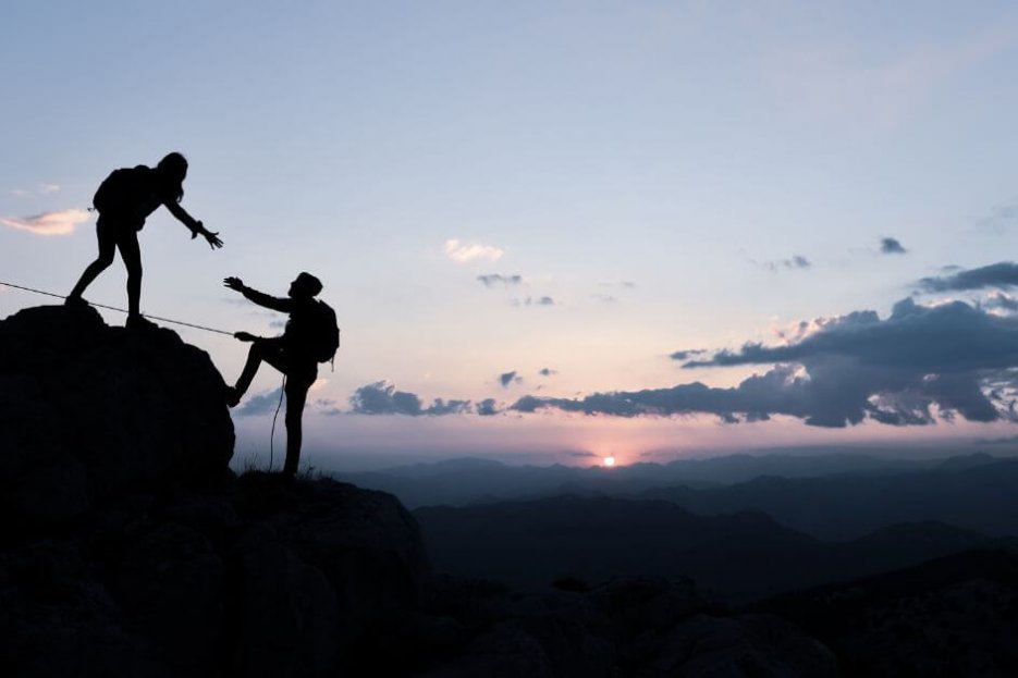 Hier siehst du zwei Kletterer auf ihrem Weg zur Bergspitze. Einer ist schon oben und reicht dem anderen seine Hand. Dieser ist kurz vorm Ziel und zusätzlich durch ein Seil gesichert. Der Hintergrund ist in blauen und grauen Tönen.