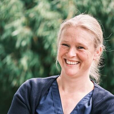 Ich stehe vor dem Bambusbusch und lache in die Kamera. Ich trage eine blaue Bluse und eine blaue Strickjacke. Mein Haar ist zu einem Knoten gebunden. Mein Haar ist dunkelblond.