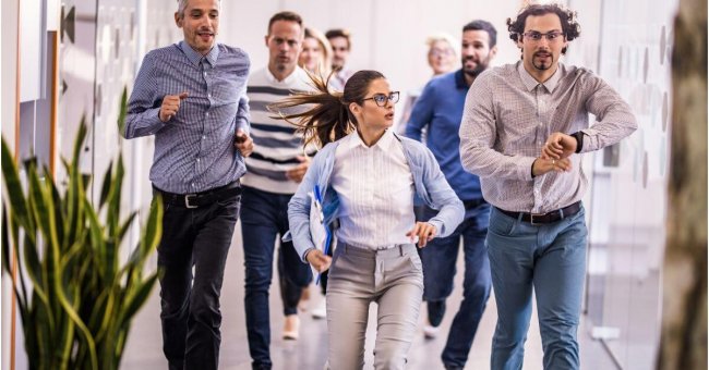 Es sind Menschen auf dem Flur zu sehen, die hektisch auf die Uhr schauen und sich beeilen. Männer und Frauen.