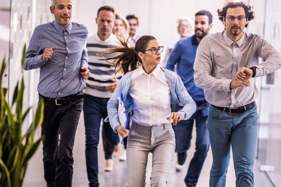 Es sind Menschen auf dem Flur zu sehen, die hektisch auf die Uhr schauen und sich beeilen. Männer und Frauen.