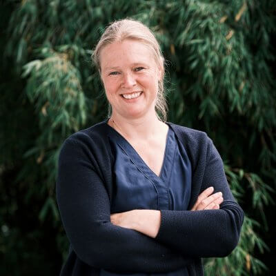 Ich stehe vor einem dunkelgrünen Bambusbusch. Mit verschränkten Armen. Ich trage eine blaue Bluse und blaue Strickjacke. Die Haare sind zu einem Knoten gebunden, ich lächle in die Kamera