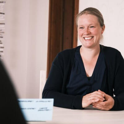 Ich sitze am Tisch, habe etwas in der Hand, links vor mir sitzt eine Person. Man kann sie nur erahnen, sie ist nicht zu sehen. Es ist ein schwarzer Ärmel zu erkennen und eine Moderationskarte. Ich lächle die Person an. Ich trage eine blaue Bluse und Strickjacke. Meine dunkelblonden Haare sind zum Knoten geflochten.