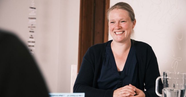 Ich bin im Coaching zu sehen mit einer anderen Person. Die zweite Person ist nur mit der rechten Schulter, von hinten zu sehen. Der Ausschnitt ist schwarz. Ich lächle und habe etwas in meinem Händen, was ich festhalte. In der rechten Bildhälfte steht ein gläserner Wasserkrug und ein Glas mit Wasser. Ich trage eine blaue Bluse und Strickjacke. In der linken Bildhälfte hängt ein Mobile von der Decke. Ich befinde mich eher im rechten Bildbereich.