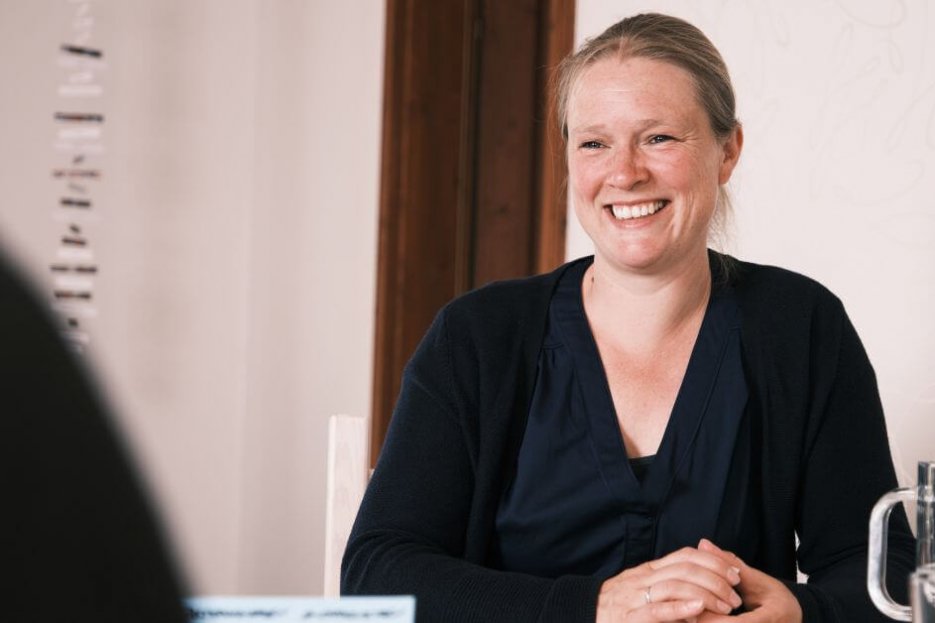 Ich bin im Coaching zu sehen mit einer anderen Person. Die zweite Person ist nur mit der rechten Schulter, von hinten zu sehen. Der Ausschnitt ist schwarz. Ich lächle und habe etwas in meinem Händen, was ich festhalte. In der rechten Bildhälfte steht ein gläserner Wasserkrug und ein Glas mit Wasser. Ich trage eine blaue Bluse und Strickjacke. In der linken Bildhälfte hängt ein Mobile von der Decke. Ich befinde mich eher im rechten Bildbereich.