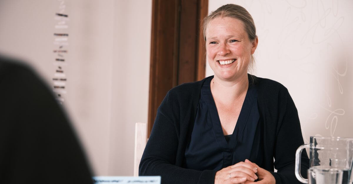 Coachinggespräch Ich bin im Coaching zu sehen mit einer anderen Person. Die zweite Person ist nur mit der rechten Schulter, von hinten zu sehen. Der Ausschnitt ist schwarz. Ich lächle und habe etwas in meinem Händen, was ich festhalte. In der rechten Bildhälfte steht ein gläserner Wasserkrug und ein Glas mit Wasser. Ich trage eine blaue Bluse und Strickjacke. In der linken Bildhälfte hängt ein Mobile von der Decke. Ich befinde mich eher im rechten Bildbereich.