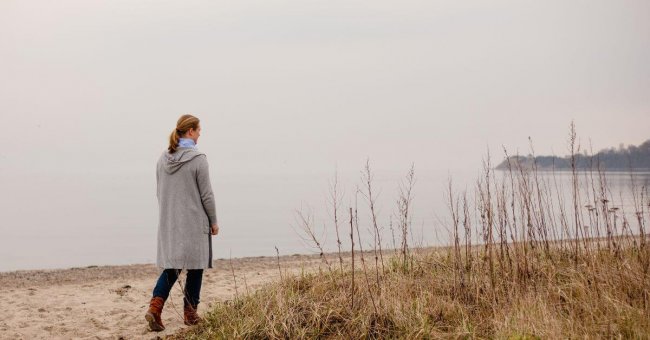Ich gehe am Strand entlang an einem bedeckten Herbsttag. Ich bin in der linken Bildseite zu sehen. Ichtrage eine graue, lange Strickjacke mit Kapuze, Bluejeans und braune Stiefel. Ich bin von hinten zu erkennen und gehe am Strand entlang. Meine dunkelblonden Haare sind zu einem Pferdeschwanz gebunden.