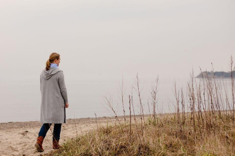 Ich gehe am Strand entlang an einem bedeckten Herbsttag. Ich bin in der linken Bildseite zu sehen. Ichtrage eine graue, lange Strickjacke mit Kapuze, Bluejeans und braune Stiefel. Ich bin von hinten zu erkennen und gehe am Strand entlang. Meine dunkelblonden Haare sind zu einem Pferdeschwanz gebunden.