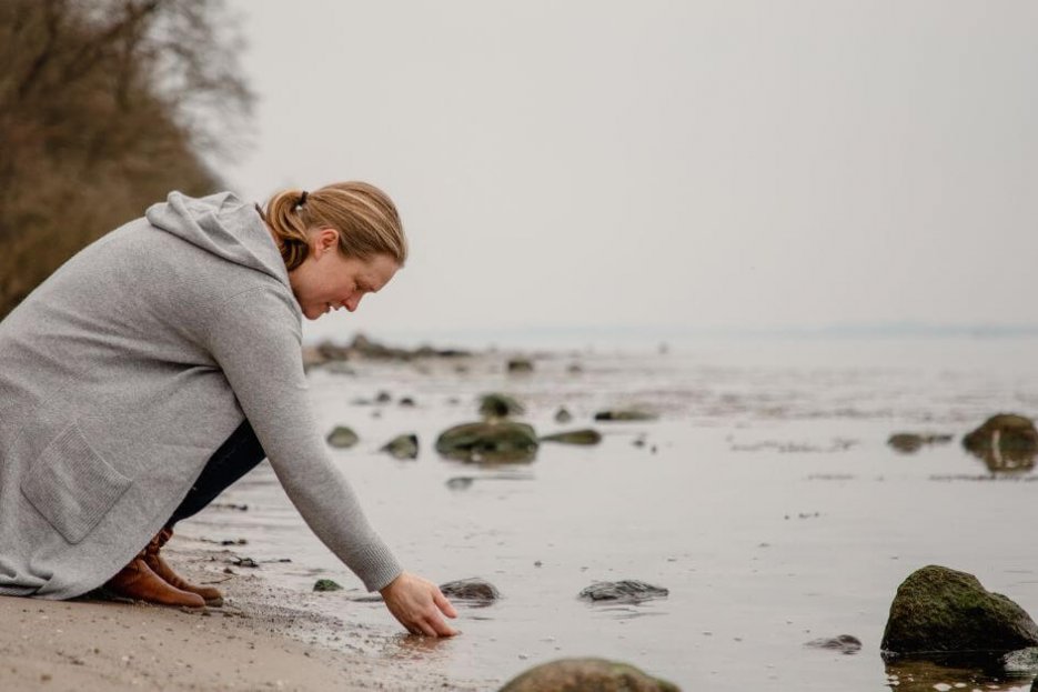 Auf dem Foto knie ich am Wassersaum der ruhigen, grauen Ostsee. Ich halte die rechte Hand ins Wasser. Ich habe eine graue. lange Strickjacke an und eine blaue Jeans. Meine Haare sind zum Zopf gebunden und reichen bis in den Nacken. Ich trage hellbraune Stiefel.