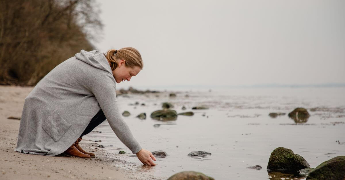 Auf dem Foto knie ich am Wassersaum der ruhigen, grauen Ostsee. Ich halte die rechte Hand ins Wasser. Ich habe eine graue. lange Strickjacke an und eine blaue Jeans. Meine Haare sind zum Zopf gebunden und reichen bis in den Nacken. Ich trage hellbraune Stiefel.