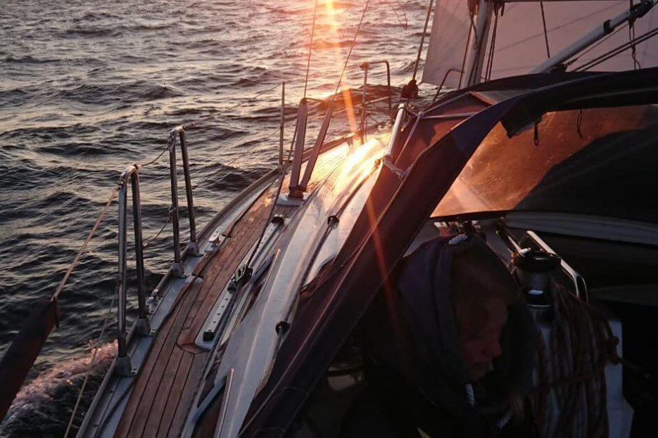 Ausschnitt eines Segelbootes. Es ist die Reeling zu erkennen und es ist zu erahnen, dass die Sonne gerade untergeht.