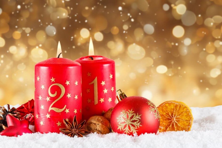 Auf diesem Foto sind zwei rote Stumpenkerzen zu erkennen. Sie sind mit goldenen Zahlen und Sternen bedruckt. Mit 1 und 2. Um sie herum liegen Weihnachtsbaumkugel und Tannenzapfen. Das gesamte Arrangement liegt in Kunstschnee. Der Hintergrund ist golden und sieht nach Lichteffekt aus.