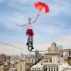Auf dem Bild fährt eine weibliche Person in luftiger Höhe mit einem Einrad auf einem Seil. Sie hat einen rot/gelben Regenschirm in der Hand. Dieser hilft ihr, die Balance zu halten.