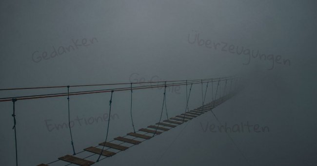 Auf diesem Foto ist eine Seilbrücke mit Holzsprossen abgebildet. Sie verschwindet auf der rechten Bildseite im Nebel. Durch den Nebel scheinen die Worte "Gedanken", "Gefühle", "Emotionen", "Überzeugungen" und "Verhaltensmuster" ganz schwach durch