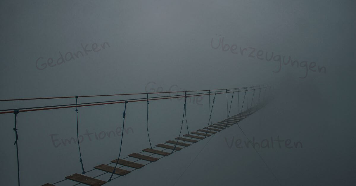 Brücke im Nebel Auf diesem Foto ist eine Seilbrücke mit Holzsprossen abgebildet. Sie verschwindet auf der rechten Bildseite im Nebel. Durch den Nebel scheinen die Worte "Gedanken", "Gefühle", "Emotionen", "Überzeugungen" und "Verhaltensmuster" ganz schwach durch