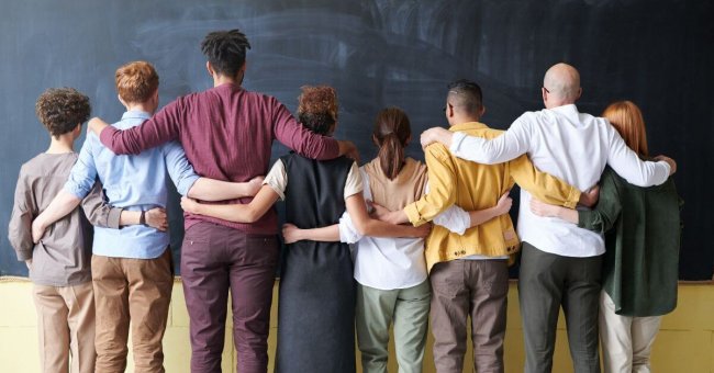 Auf diesem Foto sind acht Menschen unterschiedlicher Größe, Geschlechtes, Hautfarbe abgebildet. Sie stehen nebeneinander und haben die Arme jeweils um die nebenstehende Person gelegt. So sehen sie verbunden aus. Sie stehen mit dem Rücken zum Fotografen.