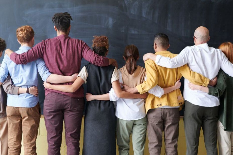 Auf diesem Foto sind acht Menschen unterschiedlicher Größe, Geschlechtes, Hautfarbe abgebildet. Sie stehen nebeneinander und haben die Arme jeweils um die nebenstehende Person gelegt. So sehen sie verbunden aus. Sie stehen mit dem Rücken zum Fotografen.