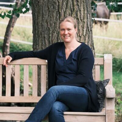 Auf diesem Foto bin ich auf einer Gartenbank sitzend abgebildet. Ich lächel in die Kamera und habe meine Beine überschlagen. Mein rechter Arm liegt auf der Rücklehne der Bank. Die Bank steht vor einen großen Baum, es ist ein dicker Stamm abgebildet. Im Hintergrund grast ein schwarzes Pferd. Ich habe eine blaue Jeans, eine blaue Bluse und Strickjacke an.