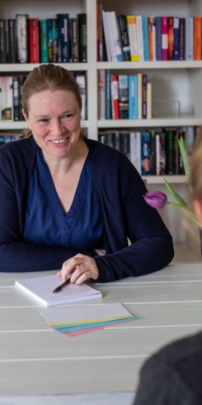 Auf diesem Bild sitze ich an einem weißen Tisch einer Klientin gegenüber. Ich habe einen Block vor mir liegen und einen Stift in der Hand. Meine Hand mit dem Stift liegt auf dem Block. Vor dem Block liegen außerdem noch verschiedenfarbige Moderationskarten. Ich lächle und habe eine blaue Bluse und eine blaue Strickjacke an. Von meiner Klientin kann man ausschließlich die linke Schulter sehen.