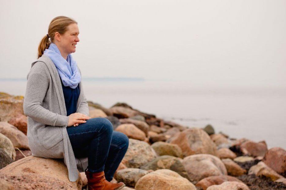 Ich sitze auf einem Stein an der Ostsee. Das Wasser ist still. Der Himmel ist bedeckt. Um den Stein herum, auf dem ich sitze, sind viele weitere Steine. Ich trage eine graue Strickjacke, hellbraune Stiefel und eine blaue Jeans. Außerdem einen hellblauen Schal.
