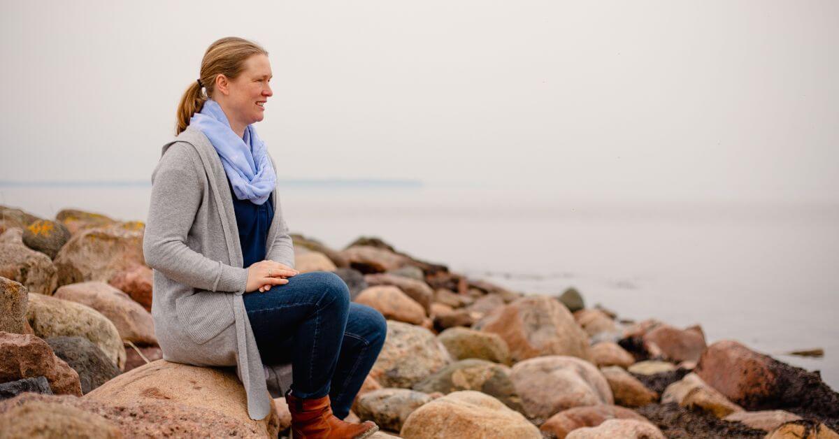 Ich sitze auf einem Stein an der Ostsee. Das Wasser ist still. Der Himmel ist bedeckt. Um den Stein herum, auf dem ich sitze, sind viele weitere Steine. Ich trage eine graue Strickjacke, hellbraune Stiefel und eine blaue Jeans. Außerdem einen hellblauen Schal.