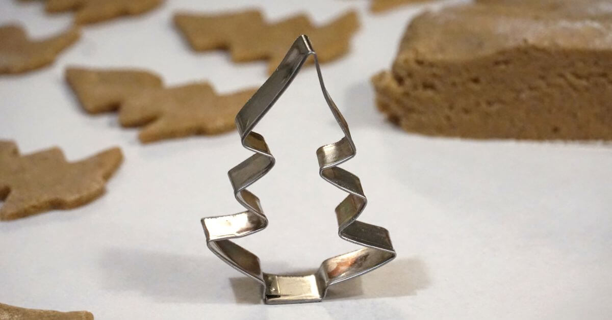 Hier ist ein verbogener Plätzchenausstecher abgebildet. Es ist eine Tannenbaum. Im Hintergrund sind bereits ausgestochene Plätzchen abgebildet und auch ein Teigrest.