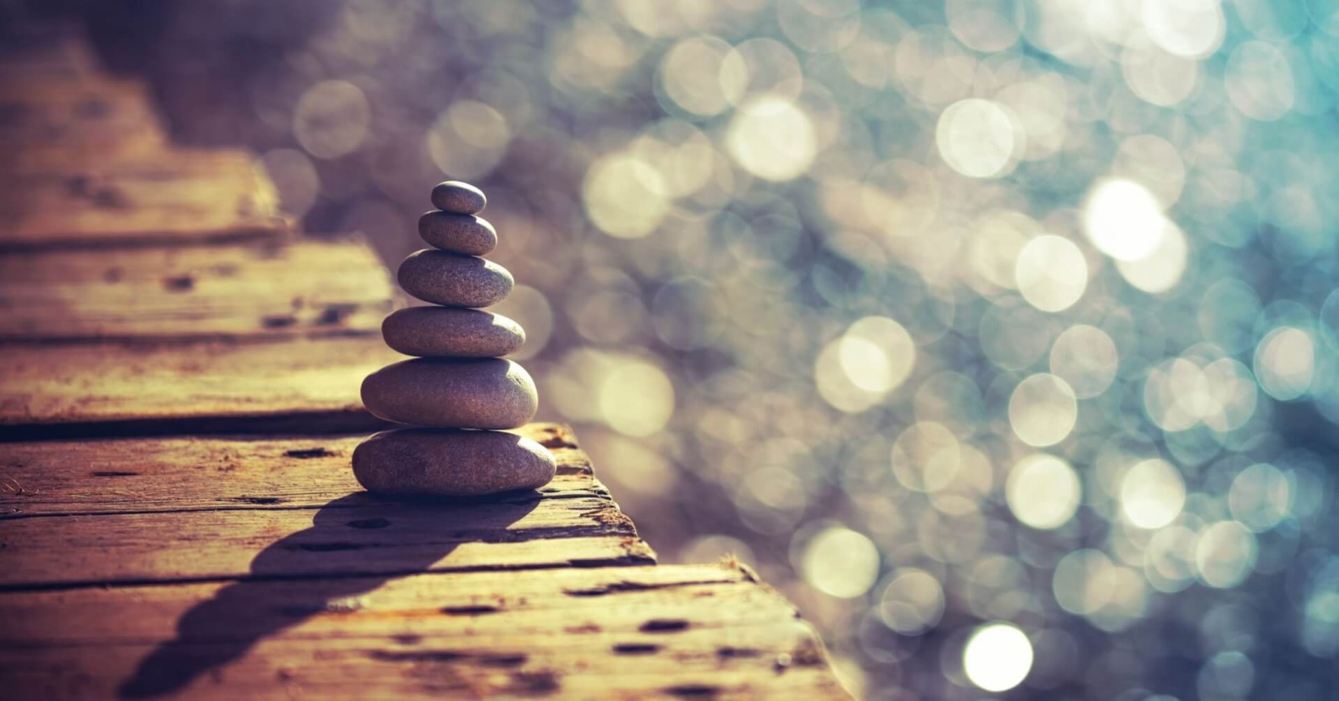 Es sind sechs Steine aufeinander gestapelt. Sie stehen auf einem braunen Holzsteg, im Hintergrund glitzert das Wasser