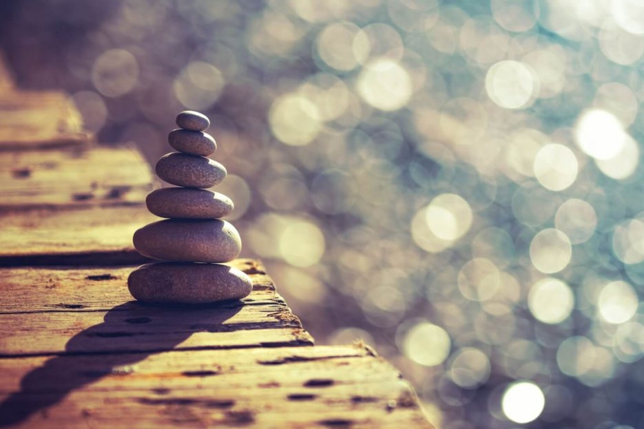 Es sind sechs Steine aufeinander gestapelt. Sie stehen auf einem braunen Holzsteg, im Hintergrund glitzert das Wasser